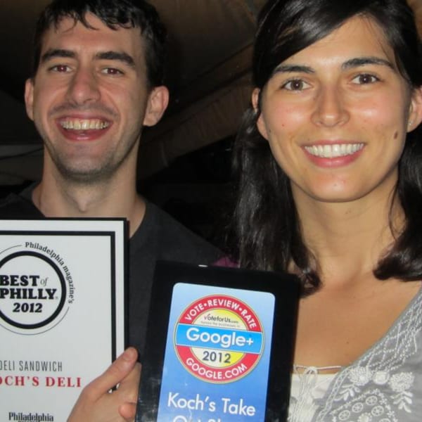 Two smiling people, a man and a woman, holding Koch's Deli awards for "Best of Philly" and Google+.