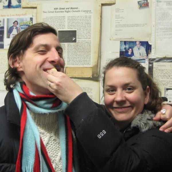 A woman feeds a smiling man a bite of food at Koch's Deli, with newspaper clippings in the background.